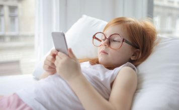 En pige med rødt hår og store briller ligger på en pude og kigger på en smartphone.