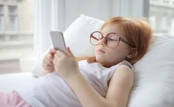 En pige med rødt hår og store briller ligger på en pude og kigger på en smartphone.