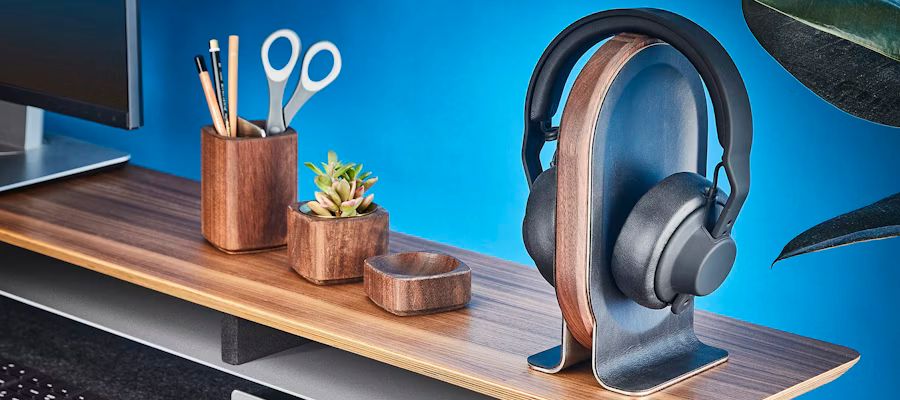 Et sort headset hænger på en elegant høretelefonholder i træ og metal, placeret på et moderne skrivebord med blå baggrund.