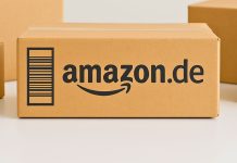 Amazon.de-pakken står centralt på et bord, omgivet af andre neutrale papkasser. Baggrunden er lys og enkel.
