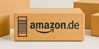 Amazon.de-pakken står centralt på et bord, omgivet af andre neutrale papkasser. Baggrunden er lys og enkel.